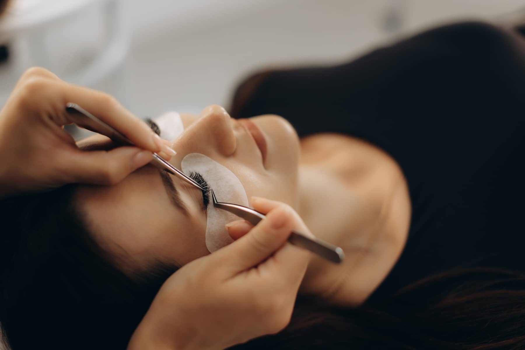 Lash technician applying individual eyelash extensions to a client lying on a treatment bed.